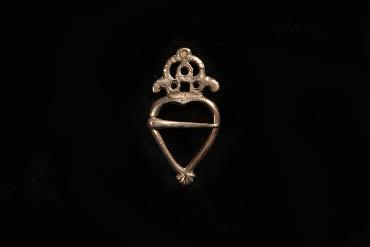En st. hjärtformad ringsölja av vitmetall med en genombruten krona upptill. Ostämplad.