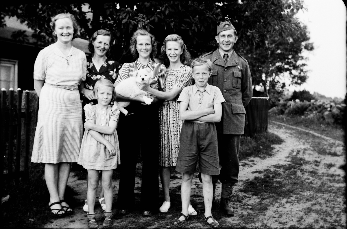 En man i uniform står tillsammans med fyra kvinnor, två barn och en vit hundvalp vid staketet till en trädgård.