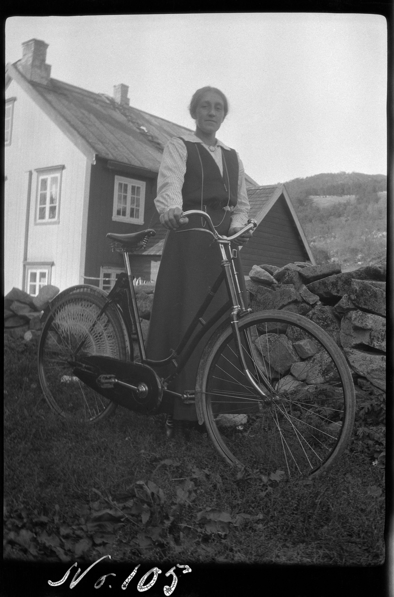 Portrett av kvinne stående med sykkel, hus og steingjerde i bakgrunnen.