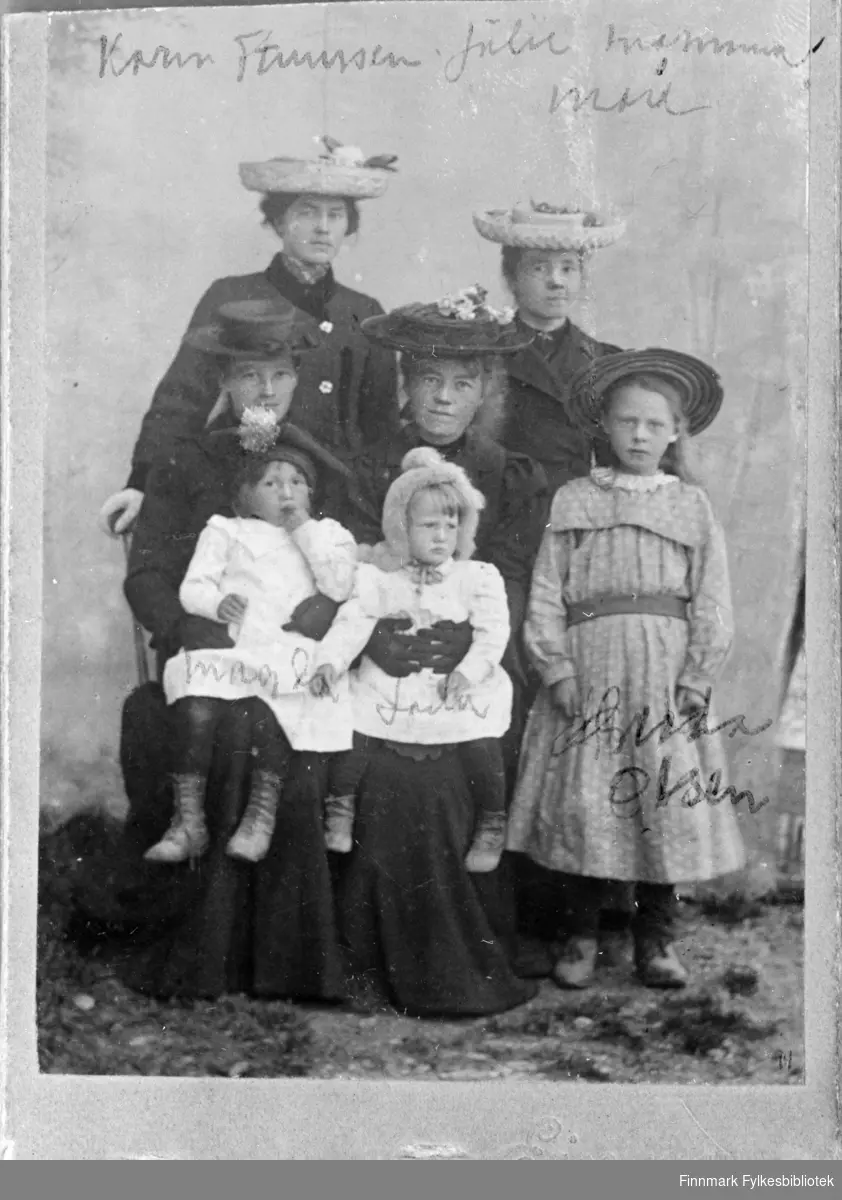Gruppebilde av fire kvinner og tre barn, oppgitt til å være tatt i Skjøtningberg. Kvinnene har datidens moteriktige stråhatter med utstoppede fugler, frukt og blomsterpynt og sorte lange kåper/kjoler. Det står navn skrevet på bildene. Det kan se ut som det står Magda (f. 1902) og Laila (f. 1903) Lund på de to små jentene i hvite kjoler. Jenta til høyre har etternavn Olsen. Over kvinnene med hatt står det muligens Karin Hanssen (?), Julie, mamma og muligens mor eller Mari(e)? . En informant har gitt en opplysning: En av damene er muligens Karen Martha Stenersen (f. 1883 i Kifjord), hun var barnepike i Skjøtningberg rundt 1900. Hun ble gift Leiknes. Fotografens navn starter på Ei... Bildet er avfotografert og vår original er et negativ av reprobildet. Bildeserien FBib.18017-001-018 har tilhørt Gerd Lund Barbala fra Friarfjord. I notatene står det at bildet er fra Skjøtningberg. Bildet ser ut til å være fotografert utendørs, det er gress og grus på bakken og det er hengt opp en tøybakgrunn bak kvinnene, muligens på en husvegg. Den minste jenta til venstre ser ut til å ha noe som ligner en samisk stjernelue på hodet, mens jenta til høyre har kyse i pels. Damene som holder barna har hansker på hendene.