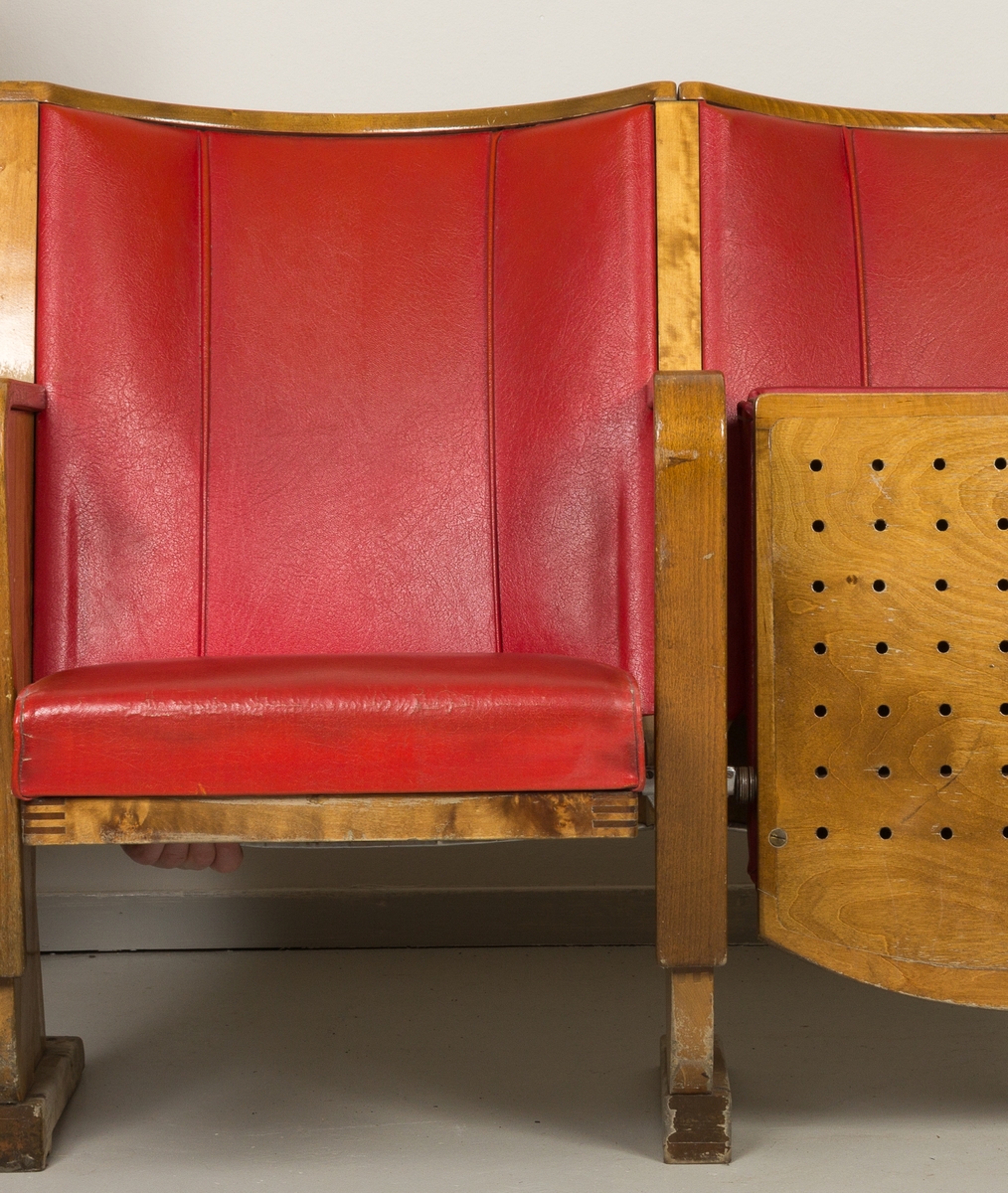 Kinostoler, to sammenhengende klappstoler i bjørk med armlener i alm, trukket i rødt skinn/skai. Brukt i Harstad kino fra 1954 til oppussingen i 1988.
