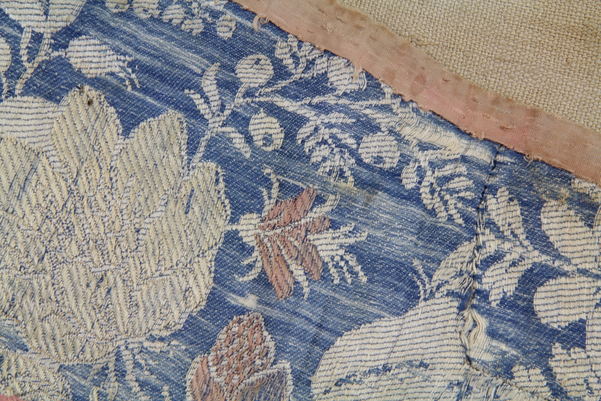 Linde av silketøy med vevd bunnmønster og brosjerte motiv. Den er foret med grovt linlerret og kantet med rosa silkebånd. Avrundet i begge ender. Sydd sammen av flere mindre stykker.