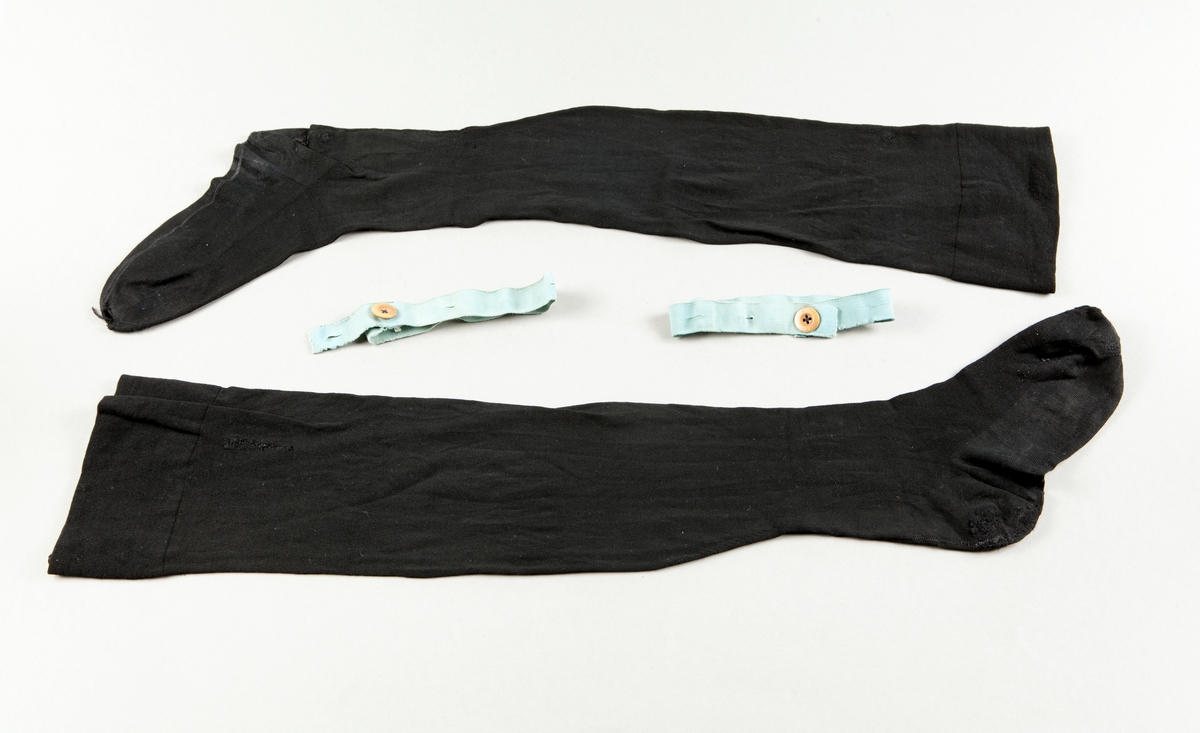 Ett par strumpor av svart ylle, maskinstickade med söm bak. Strumpeband av blått resårband med brun knapp. Text: Deusch Mode.
Stoppar och hål på häl och tå.