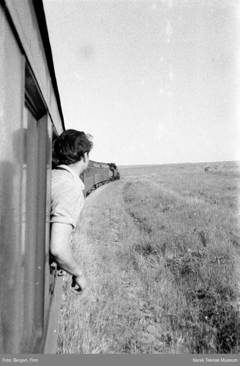 Person som lener seg ut av vinduet på et tog. Flatt, øde landskap, strekningen Istanbul - Bagdad, Taurus Express.
