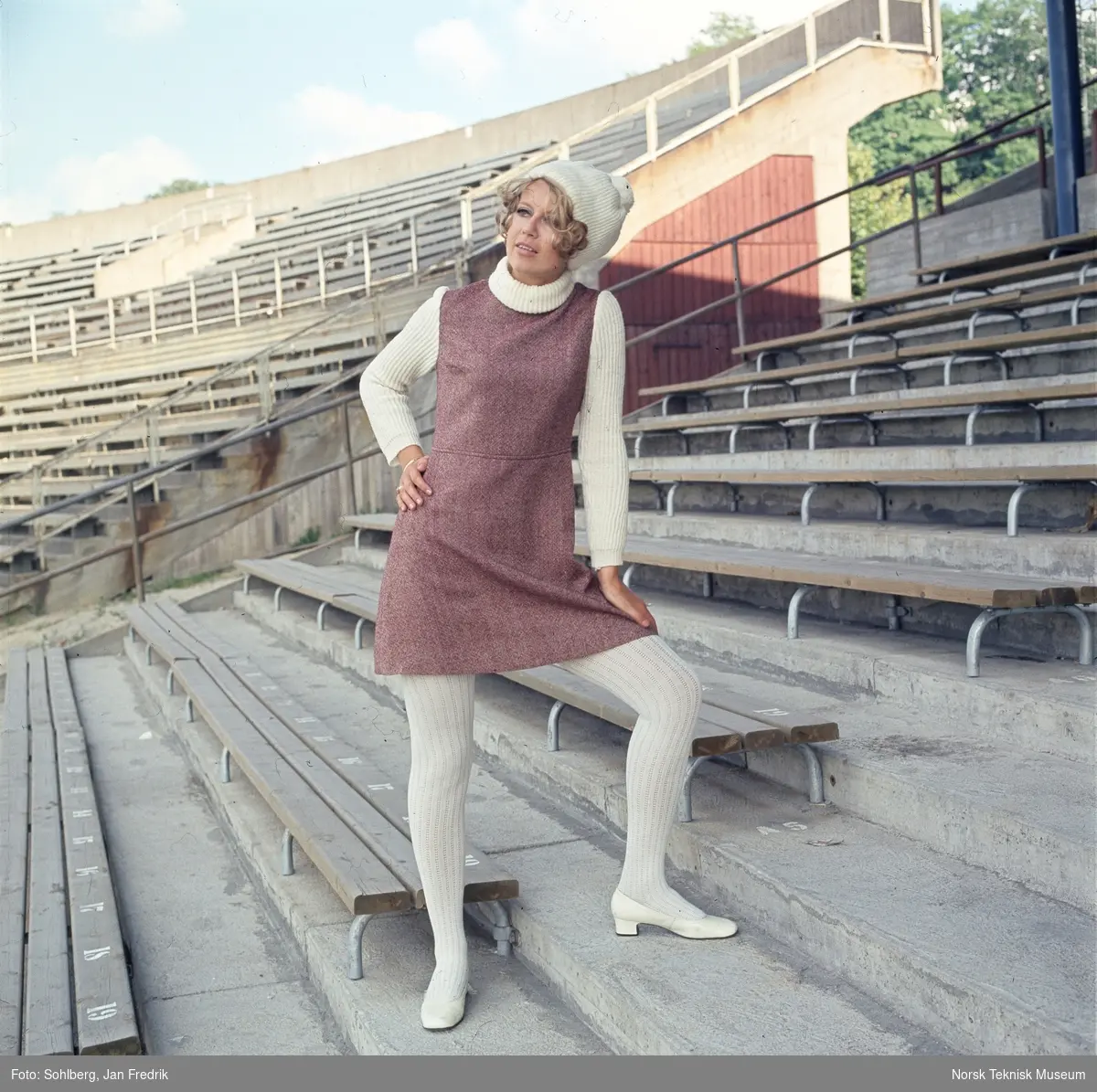 Motebilde, kvinne i lilla ullkjole med hvit genser, sko, strømper og alpelue, på tribunen på idrettsarena. Bildet er en del av en serie der noen er publisert i ukebladet Det Nye 1969, nr. 38.
