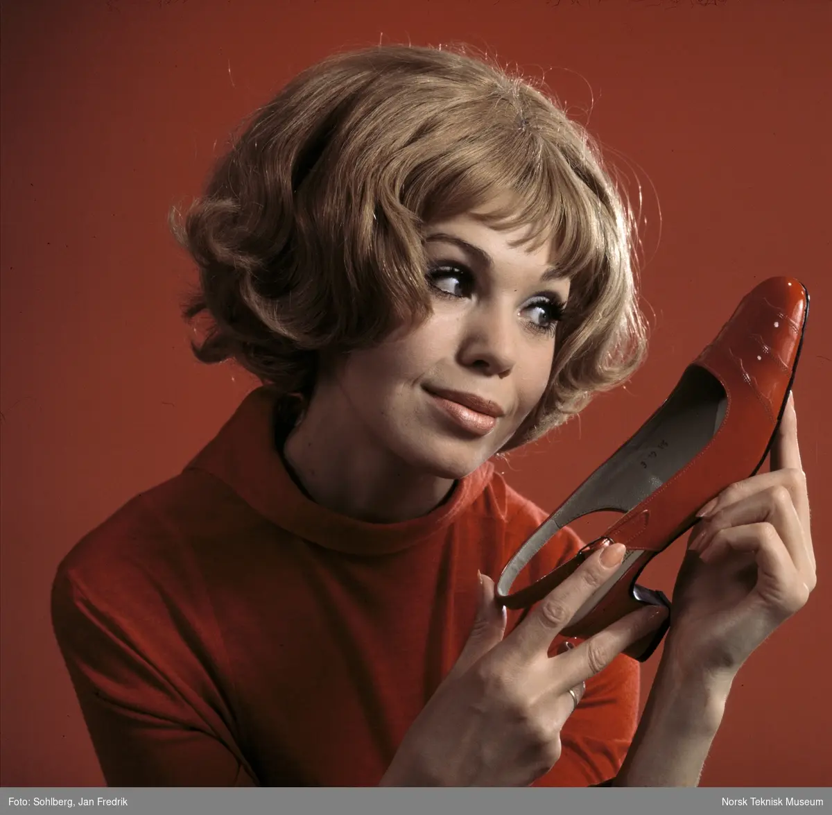 Tidlig norsk reklame- / motefoto. En kvinnelig modell holder opp en rød sko med høy hæl, hun virker fornøyd med tidens skomote. Hun har rød bluse. Rød bakgrunn.
