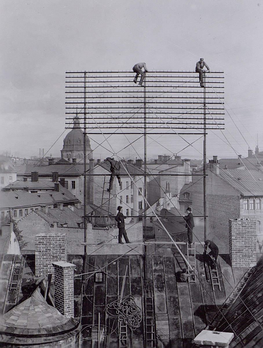 Ledningsbygge. Arbete i elefongalge på taket till Biblioteksgatan 10, Stockholm. 1890-talet (troligen 1898 enl takkontrakt). (Hedvig Eleonora Kyrka i bakgrunden). På landgången syns till vänster ingenjör K.W. Gerdhem och till höger linjebyggmästare C E Nilsson.