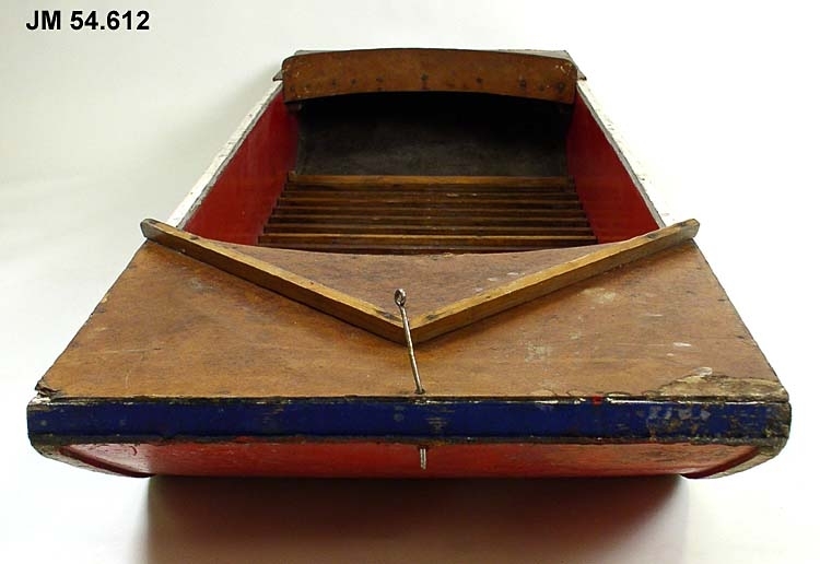 Barnbåt av masonit och trä med flat botten och låga kanter. Den är blåmålad på utsidan och rödmålad på insidan. Botten inuti båten täcks av träribbor. Båten är täckt i för och akter. En metallpinne med ögla sitter i fören för att förankra båten med. Denna kunde även användas för att fästa seglet i. I ett hål i båtens bakre ände har troligtvis en liknande ögla suttit, för att fästa bakre delen av seglet i. Mitt i båten ställdes en mast ned. Undersidan är rödmålad.