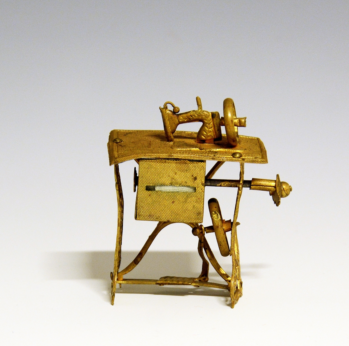 Liten bordsymaskin i miniatyr. Utført i gult metall, noe skjev i enkeltdelene.