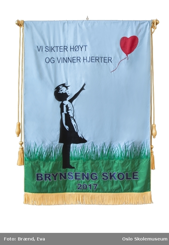 En barneskikkelse stående på gress som holder hånden opp mot en rød hjerteformet ballong. Øverst på fanen står skolens visjon (se Andre opplysninger). Nederst på fanen står skolens navn og året den ble etablert.
