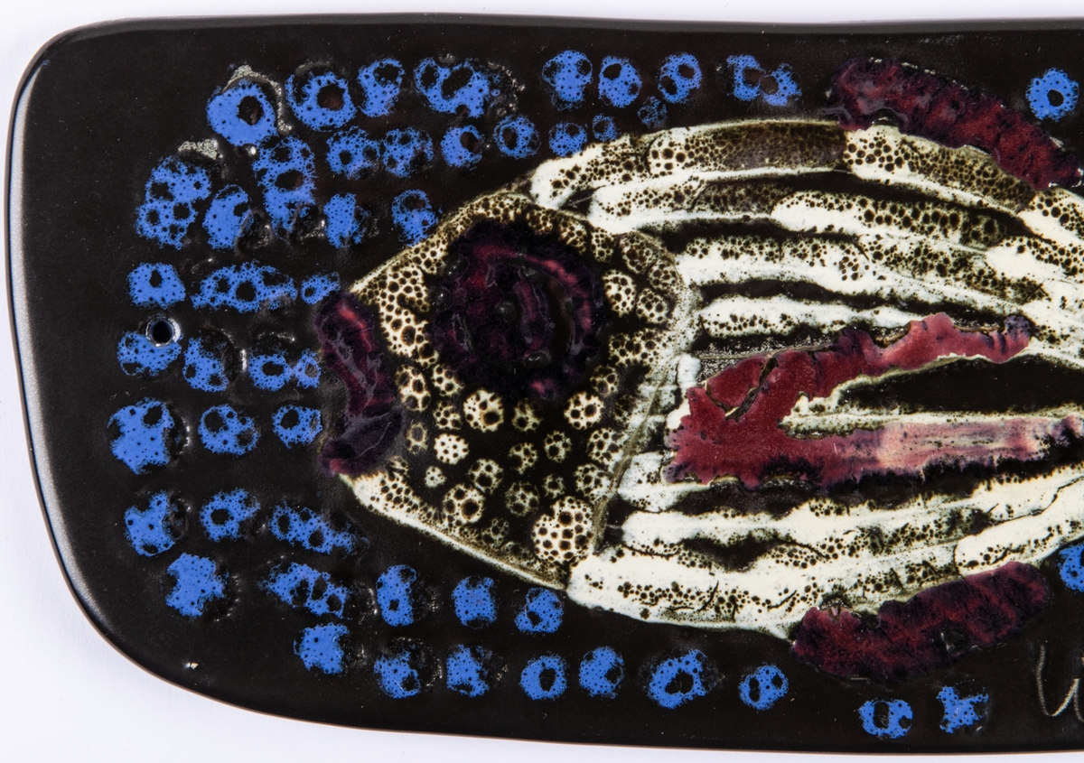 Väggplatta, flintgods, dekor av fisk. Glasyr Mangania med blått, vitt och rött mönster. Formgiven av Lillemor Mannerheim.