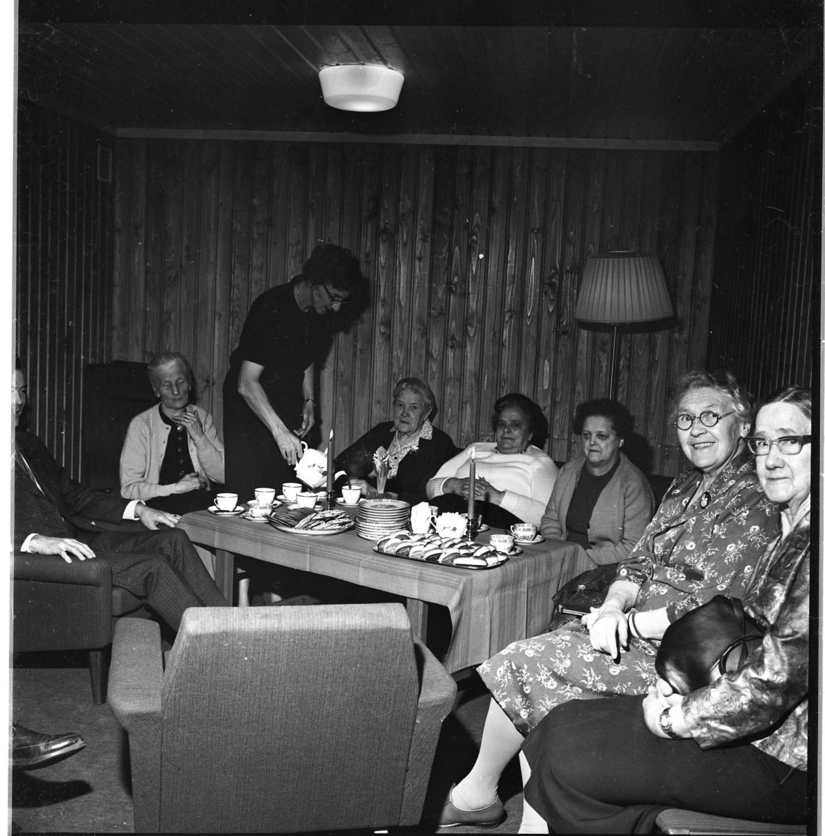 Äldre damer sitter i fåtöljer och soffa omkring ett rektangulärt bord med duk, tända ljus samt kaffekoppar och kakfat. En yngre kvinna, Karin Holm, häller upp kaffe till Ida Svensson.