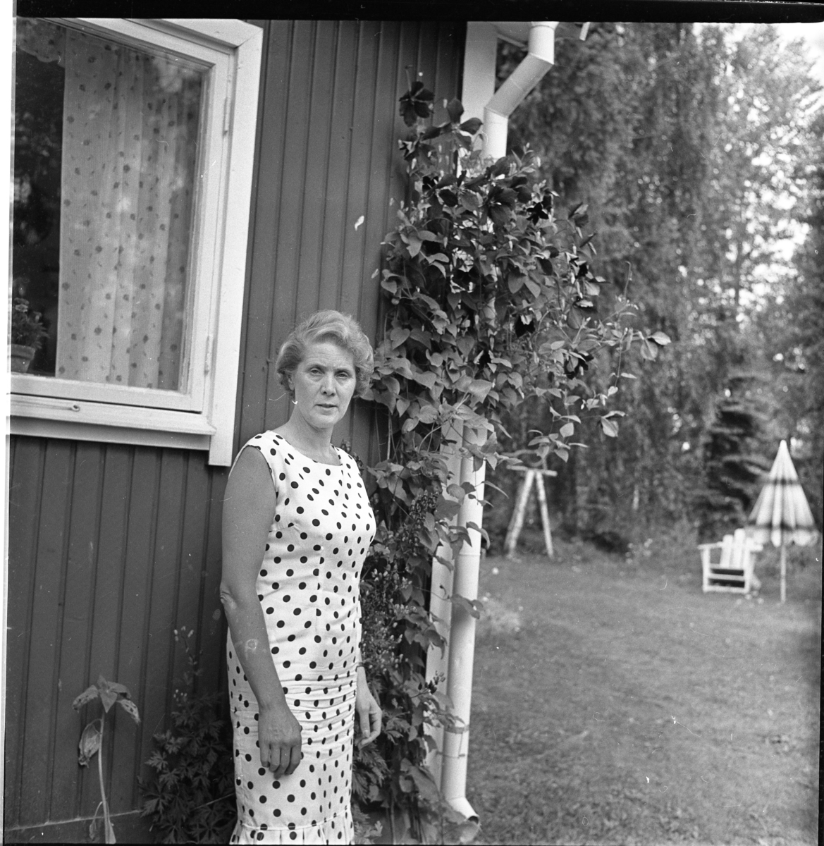 Birgit Heldebrandt i prickig klänning står vid en husvägg, möjligen en sommarstuga. Det är sommar.