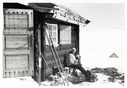 Kvinne sitter langs husvegg ved hytte. Ski står langs husveg