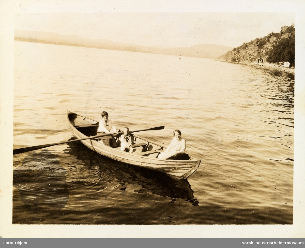 To kvinner og ett guttebarn iført sommerklær sitter i robåt på vann