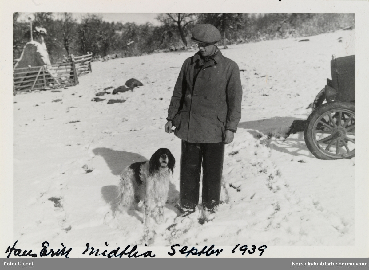 Hans Erik og hund står i snøen. Ved siden sees forhjulene til et kjøretøy, bak sees inngjerding