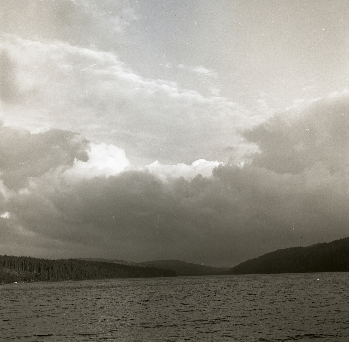 Böljande vatten bland skog, berg och en himmel med moln. Törn 1989.