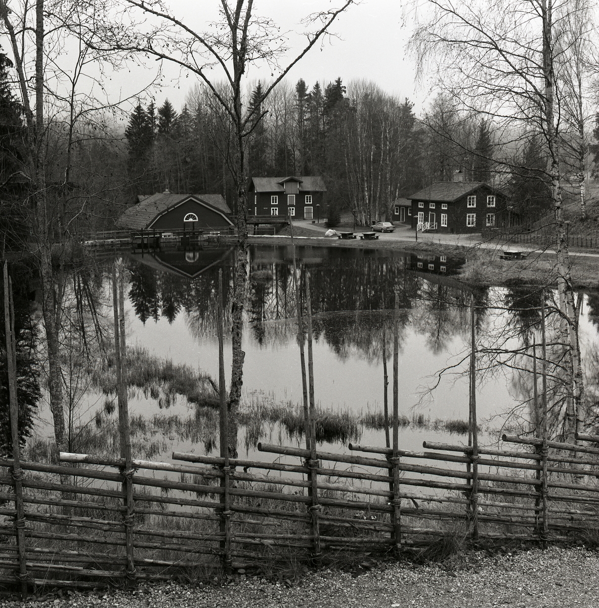 Vid spegelblankt vatten finns några byggnader med skogen bakom. Det finns en grusväg, vass i vattnet, träd, gräs, gärdesgård och himmel. Trolldalen 1990.
