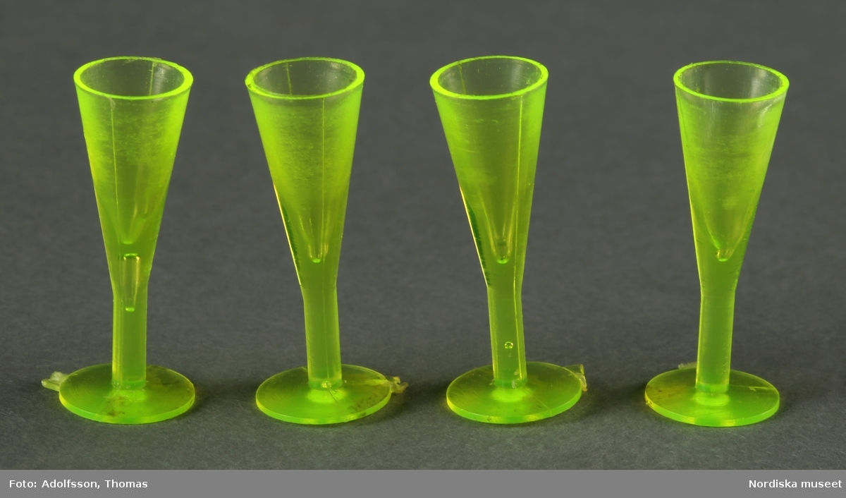 Fyra neongröna champagneglas av plast. Hör till det uppdukade matbordet (NM.0333310 a) mitt i matsalen i dockskåpet NM.0331721+.