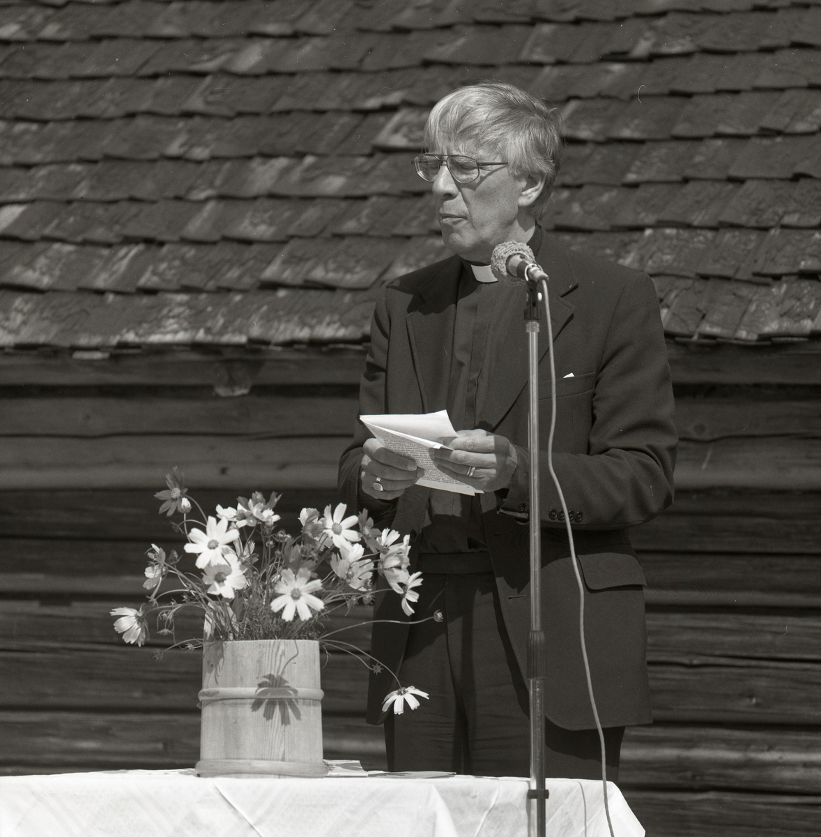 En man klädd i prästkläder håller ett tal vid mikrofonen. Bordet har en vit duk och en kruka av trä med blommor i. Bakom är en vägg i trä och tak av tegel. Harsa 5 augusti 1990.
