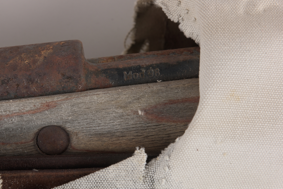 Gevær med trestokk, sluttstykke og bærestropp. Det er rester av et lerrets omslag med lærforsterkninger. Det kan være et hylster tilpasset våpen. Treverket er værbit og metalldelene er rusten av å ha ligget ute i mange år.