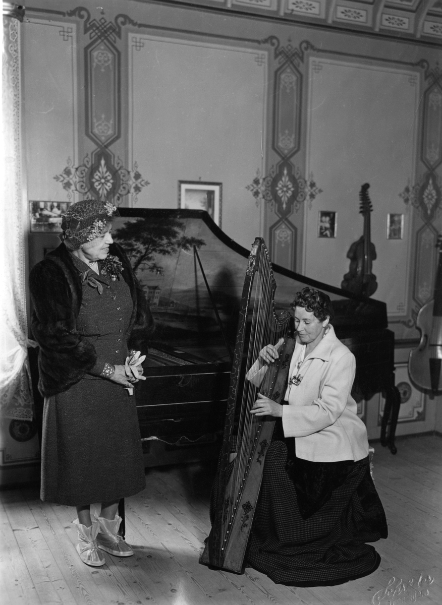 Bilde av en skotsk harpistinne og en amerikansk komponist i Mozartsalen. Harpistinnen Liss Russel Ferguson spiller på harpen og Mrs. Clara Edwards, komponisten, står ved siden av.  Begge er på sightseeing på Ringve som hurtigrute-passasjerer.