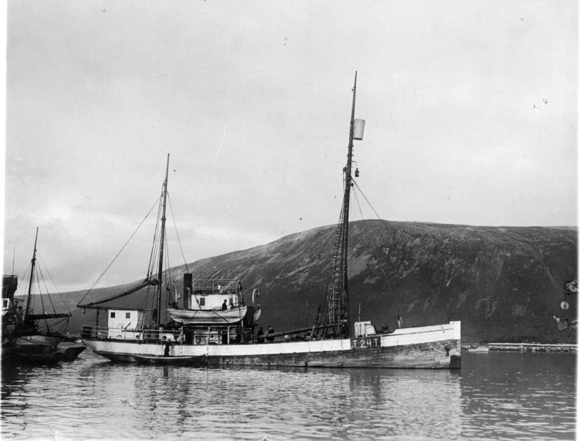 Ett norskt fångstfartyg med beteckningen T 24 T. En utkikstunna i toppen av främre mast. En eka med en stående man i ligger intill fartyget.