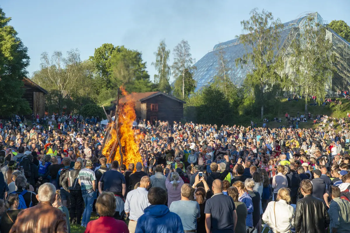 Sankthansbålet brenner midt på en slette fylt med glade tilskuere, de gamle husene på Hedmarkstunet og Hamardomen vises i bakgrunnen.