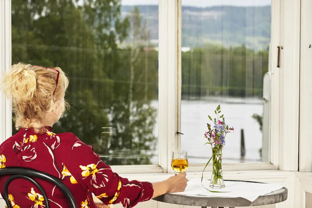 Dame i rød bluse som med ryggen til. Ser ut av vinduet ut mot elva.  Damen sitter på en stol og holder høyre hånd rundt et glass med eplemost som er plassert på et bord. En glassblomstervase med blomster