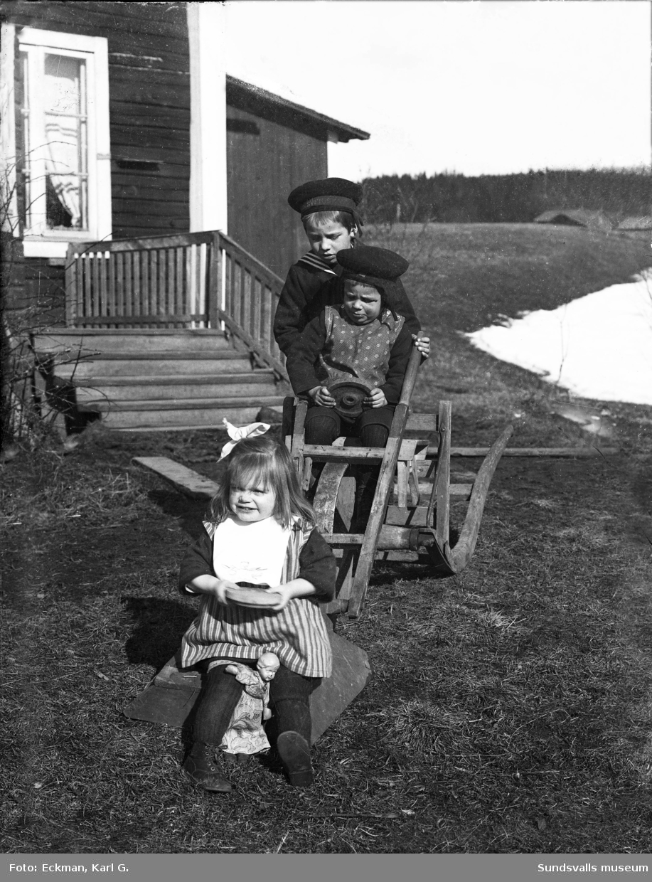Barnen Eckman (Robert, John och Rakel) leker med några skottkärror som om det vore en bil. I bakgrunden ses bostadhuset och utbyggnaden där fotoateljén fanns inhyst. Ur fotograf Karl Eckmans samling.