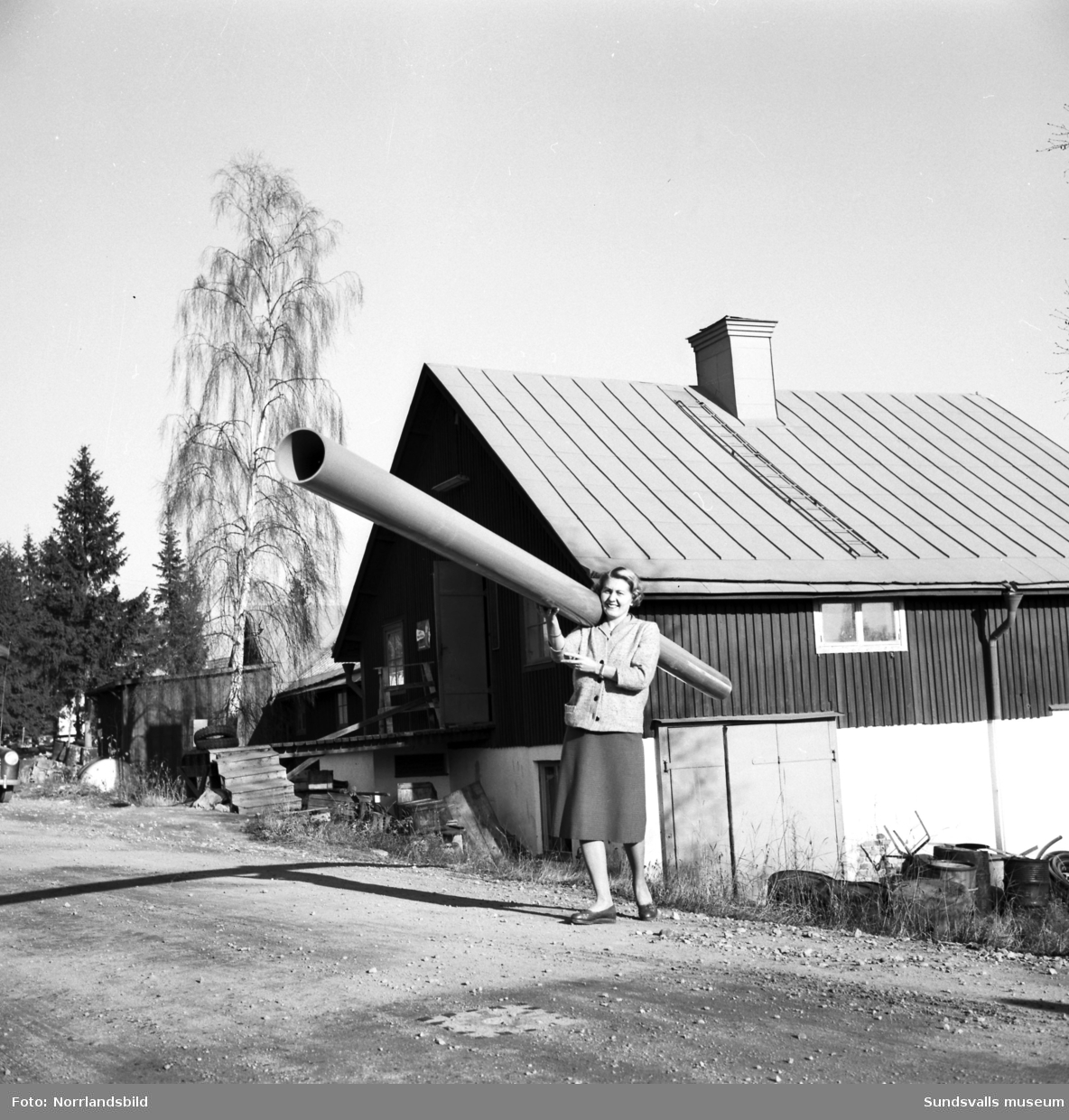 Firma Bertil Ericsson, Sundsvall (senare AB Centrifugalplast). En kvinna håller ett stort plaströr på axeln utanför en lantligt belägen lagerlokal.