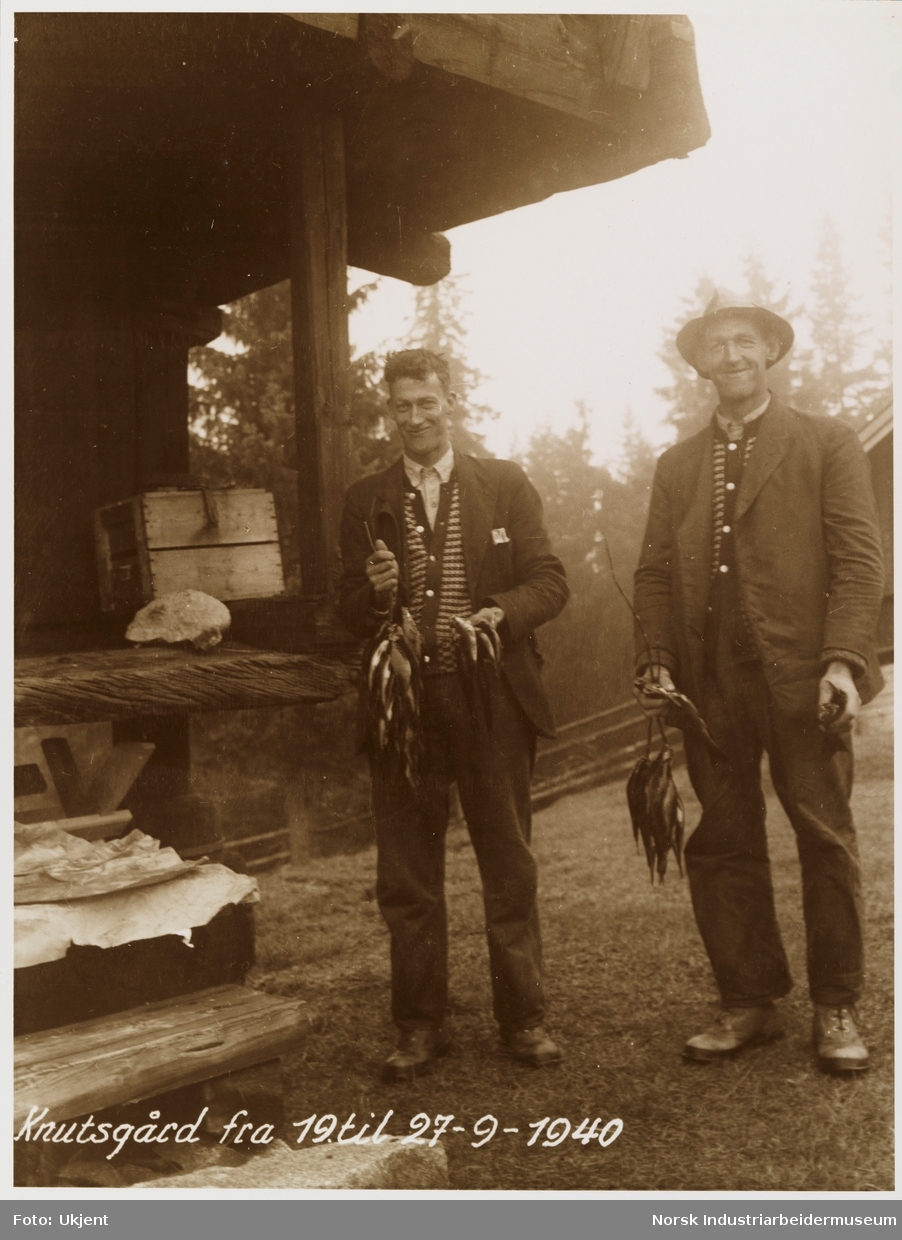 To menn foran loft på Knutsgard smiler og holder fisk i hendene