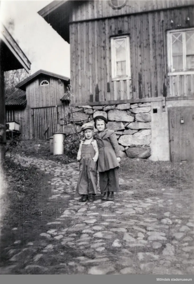 Porträtt av två små barn på gården Christinedal i östra Mölndal på 1950-talet. I bakgrunden ses ett par av gårdens ekonomibyggnader. Gården har gett namn åt området Kristinedal och var belägen i vinkeln mellan nuvarande Rådavägen och Brovaktaregatan. År 1960 såldes gården till Fastighetsbolaget Betongblandaren (Skånska Cementgjuteriet) och revs när det nuvarande bostadsområdet anlades.