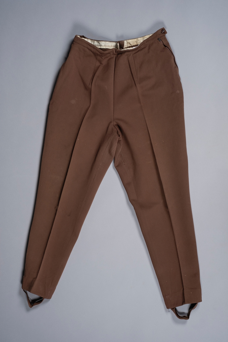 Bukse i brunt stoff som smalner nedover og ender med fotløkker. Buksen lukkes med glidelås i siden. På den andre siden er det en glidelåslomme.