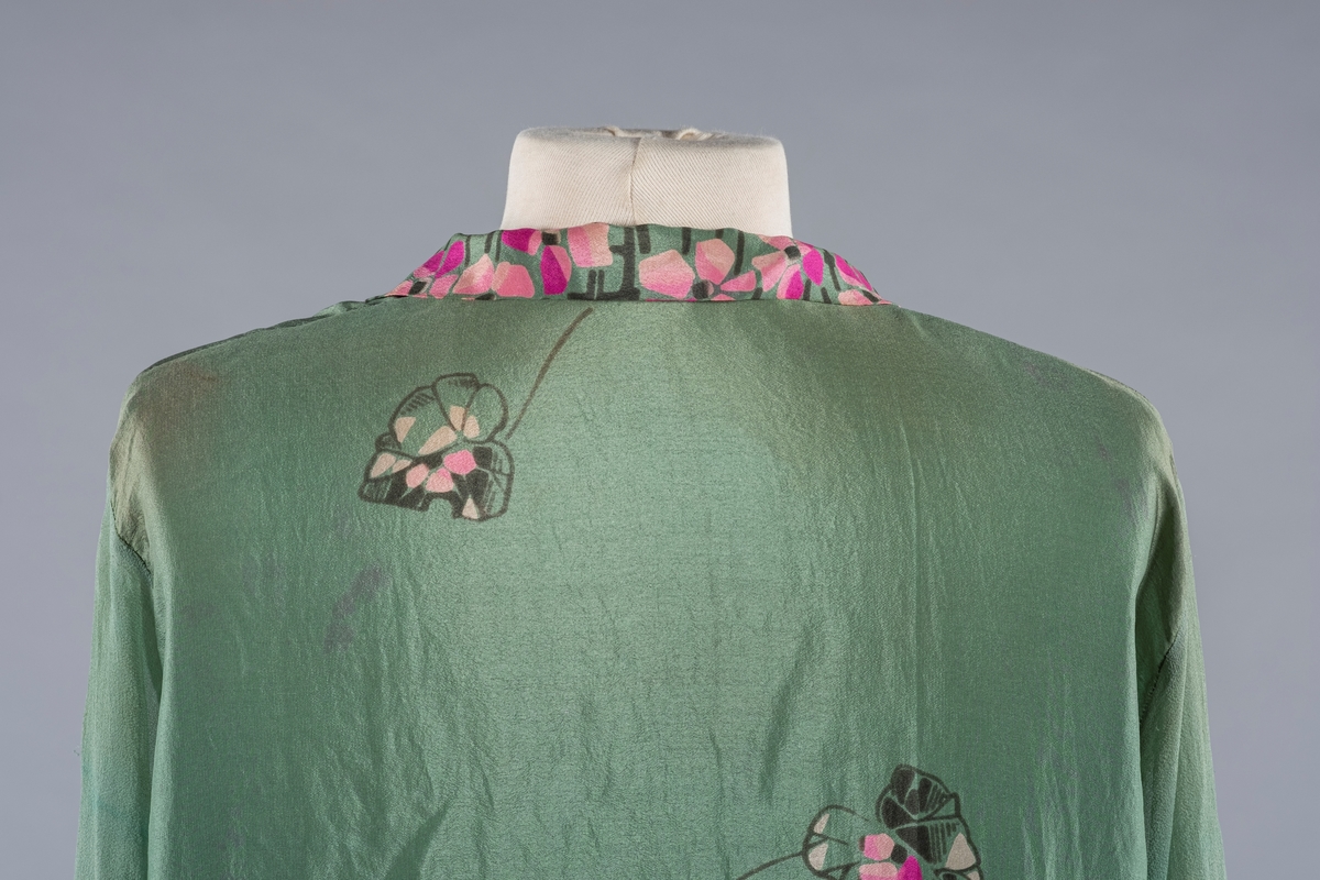 En grønn silkekjole med rosa blomstermønster i Charleston-fasong. Den har lange ermer og V-ringet halsåpning. Ermene er i krepp.chiffon. Mansjettene er smale med trykknapper. Det er svarte stofftrukne pynteknapper i halsåpningen. På sidene er stoffet rynket med påsydd spensel og 2 stofftrukne pynteknapper i sort.