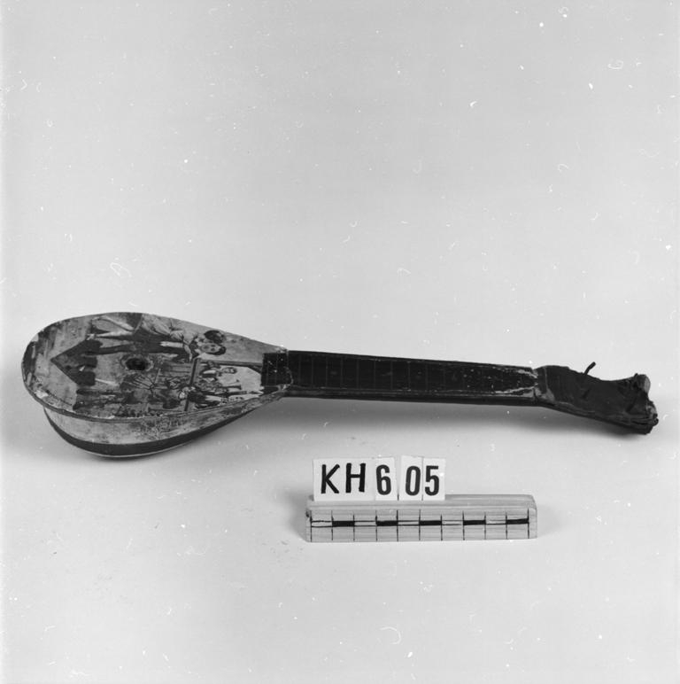 Mandolinliknande instrument, dekorerat med ett dansande par till en blåsorkester. Strängar saknas.
Märkt: "Made in Czechoslovakia".