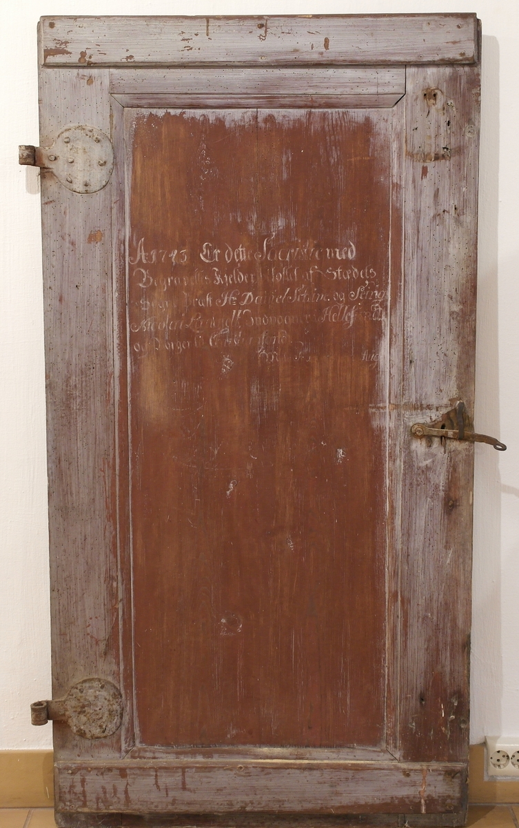 Dør i furu med smidde jernbeslag og hengsler. Døra er opprinnelig rødmalt, og malt grå over i ettertid. På dørspeilet er det malt en tekst.