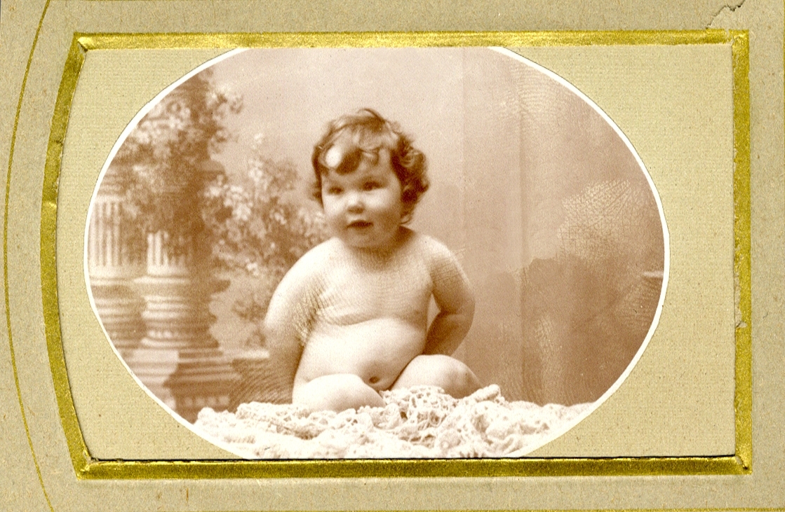 Foto av en liten naken baby som sitter på en fäll. 
Helfigur. Ateljéfoto.