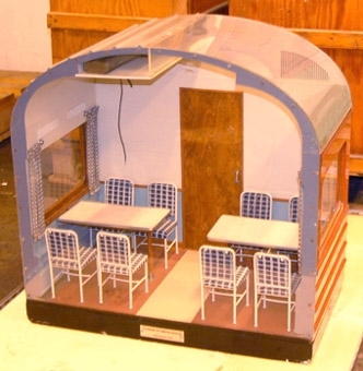Modell i skala 1:5 av en sektion av en Ro3 restaurangvagn. Inredningen har brun och beige golvbeläggning, samt grå väggar och tak. 
Brun dörr, och brun- och vitrandiga gardiner. 
2 bord med perstorpsplatta, med 8 stolar med sits och ryggstöd klädda med blått och vitt rutigt tyg.

Modellen står i en specialbyggd transportlåda.