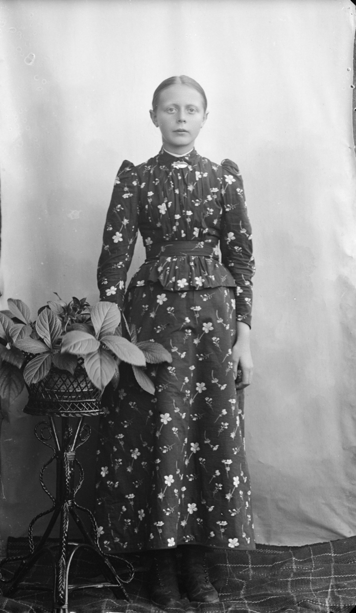 Kvinne som står ved siden av et rottingbord med blomster. Hun har glatt hår med midtskill, kledd i lang stakk og overdel med puffermer i blomstrett stoff