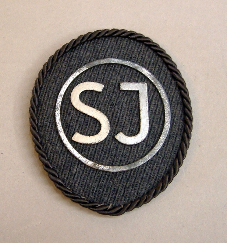 Märke för uniformsmössa för SJ bussförare. Ovalt märke av grått tyg och en grå kantsnodd. "SJ" samt en ring runtom av metall.