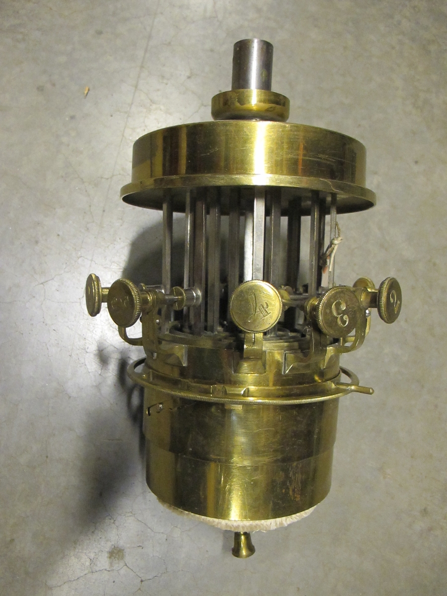 Lampnäbb, 6-veksbrännare till mekanisk lampa för oföränderlig oljeyta (för konstant nivå) och fotogen. 6 st med rundveke försedda koncentriska dubbla rör med luftmellanrum. Vekarna fästade vid en med kuggstång försedd ring. Kuggstängerna löper hermetiskt i rör med rektangulär genomskärning vars nedre är anslutna till en liten cylindrisk oljebehållare varifrån oljan leds upp till vekarna. Förbindelsen mellan den mindre behållaren och en större behållare, anordnad för oförändrad oljeyta, sker med kvicksilverlås (rörtappen sticks ned i en med dubbla väggar försedd skål, vars mellanrum innehåller kvicksilver). Numrerade ställskruvar, en till vardera av de två minsta vekarna, två till vardera av de fyra största. Samtliga skruvar anslutna till kuggstångsrören med packningsboxar för att förhindra läckage. Lampglaset vilar på en höj- och sänkbar ring som omsluter brännaren. Brännaren vilar på ett stativ som ej tillhört lampan.