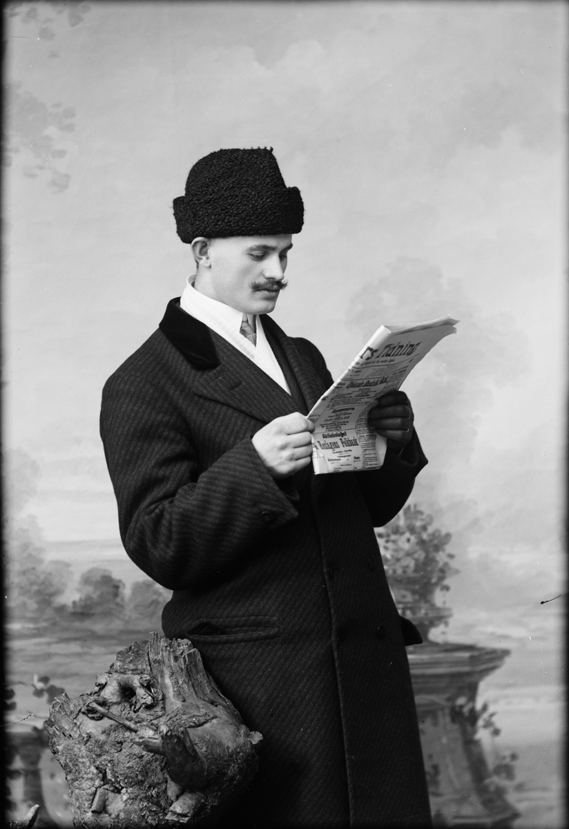 Ateljéporträtt - man i ytterkläder står och läser tidning, Östhammar, Uppland