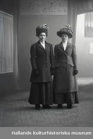 Ateljébild. Två kvinnor i helfigur. Båda bär ytterkläder och hattar. Beställare: Gerda Persson.