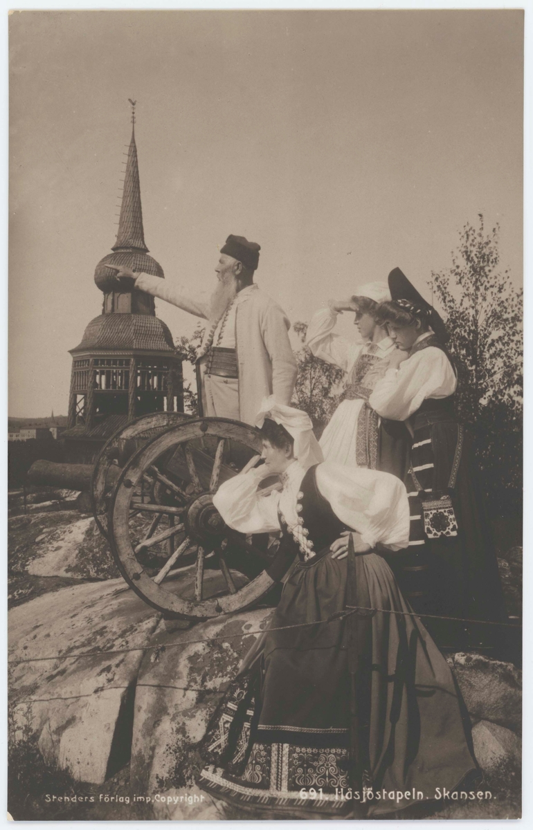 Vykort med motiv från Skansen. Tre folkdräktsklädda kvinnor och en man vid kanon. I bakgrunden Håsjöstapeln.