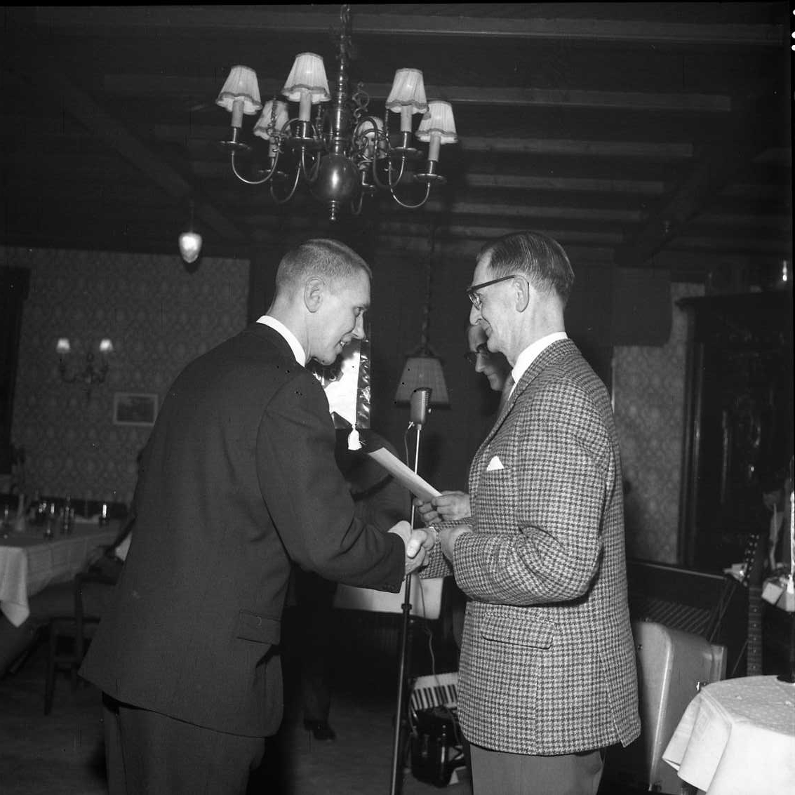 Två män handhälsar, en yngre i mörk kostym med slips och en äldre - Bertil Ander - i rutig blazer och udda byxa. Det är utdelning av utmärkelser i samband med årsmöte för Gränna AIS.