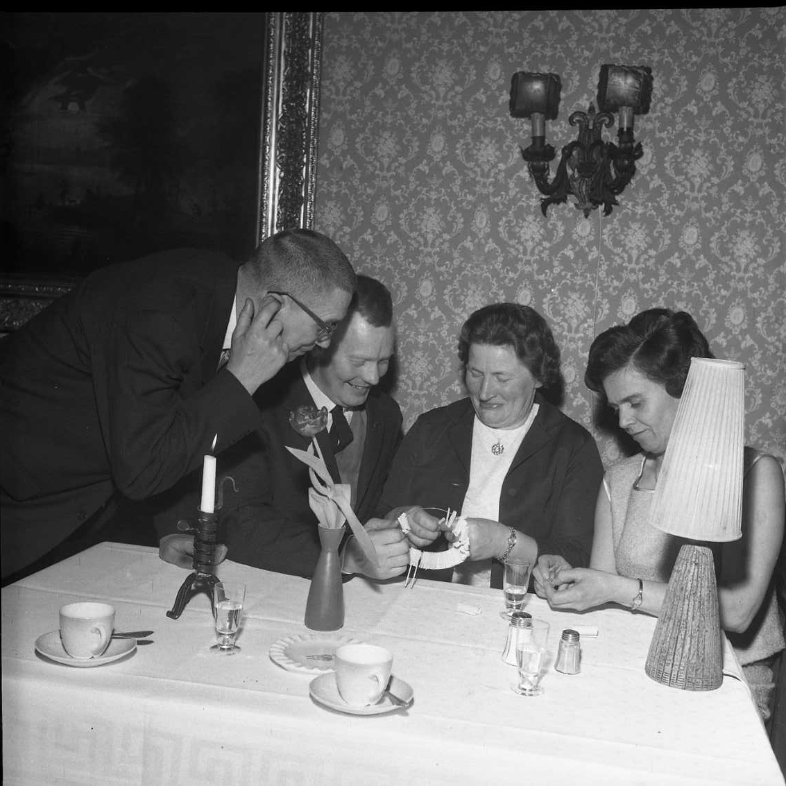 Två festklädda skrattande kvinnor sitter vid ett bord med kaffekoppar, bordslampa och tänt ljus. De köper och rullar upp lotter som de köper av en man som sitter intill dem till vänster. En andra man står lutad över dem från samma sida. Han håller ett finger över höger öra som för att höra bättre.