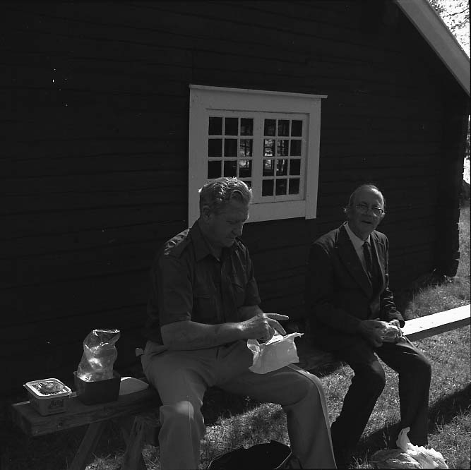 Två män, Larsson och Harry Linder i glasögon till höger, sitter på en bänk vid en husgavel till ett äldre torp och fikar. Det är sommar och de sitter i skuggan.