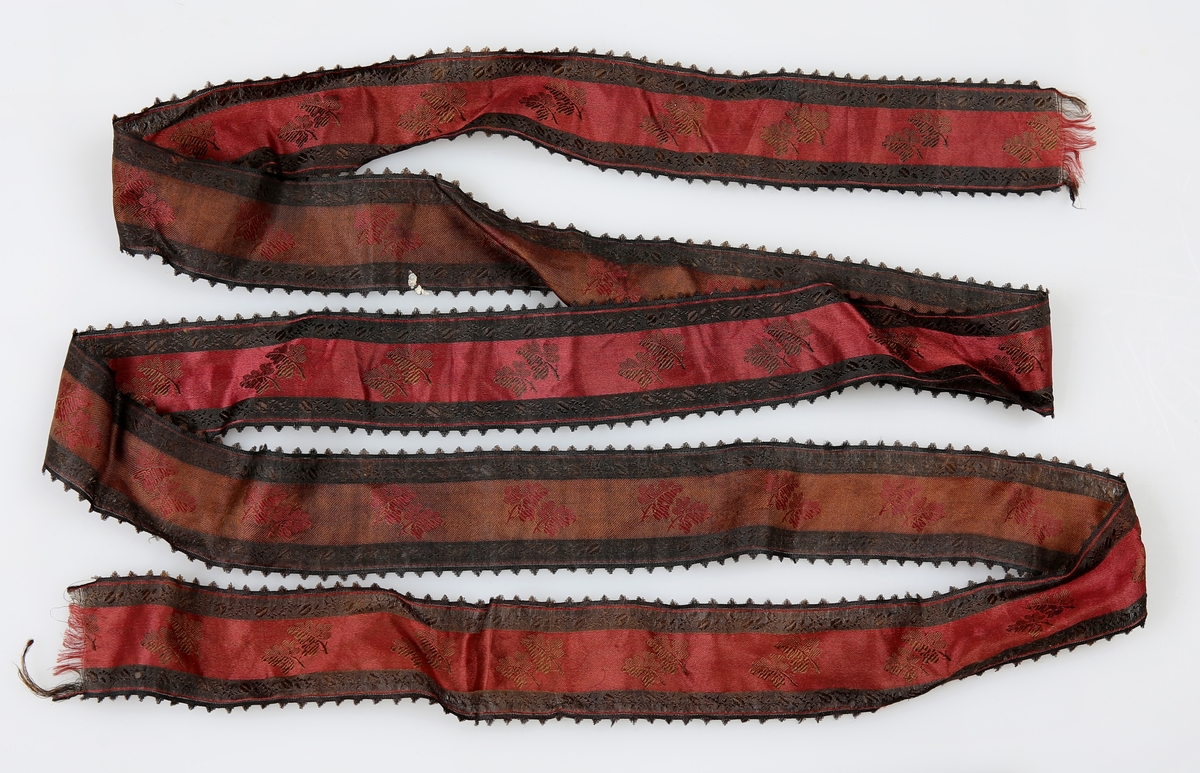 Silkebånd i svart og rødt. damaskvevd. Pigoter ut på hver side av båndet. 