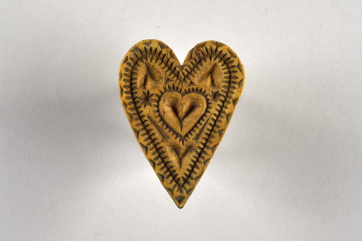 Hjärtformad kakstämpel av trä. Stämpelsidan dekorerad med karvsnittsdekor i form av hjärtan, stjärnor, trianglar och streck. Skuren knopp på undersidan.